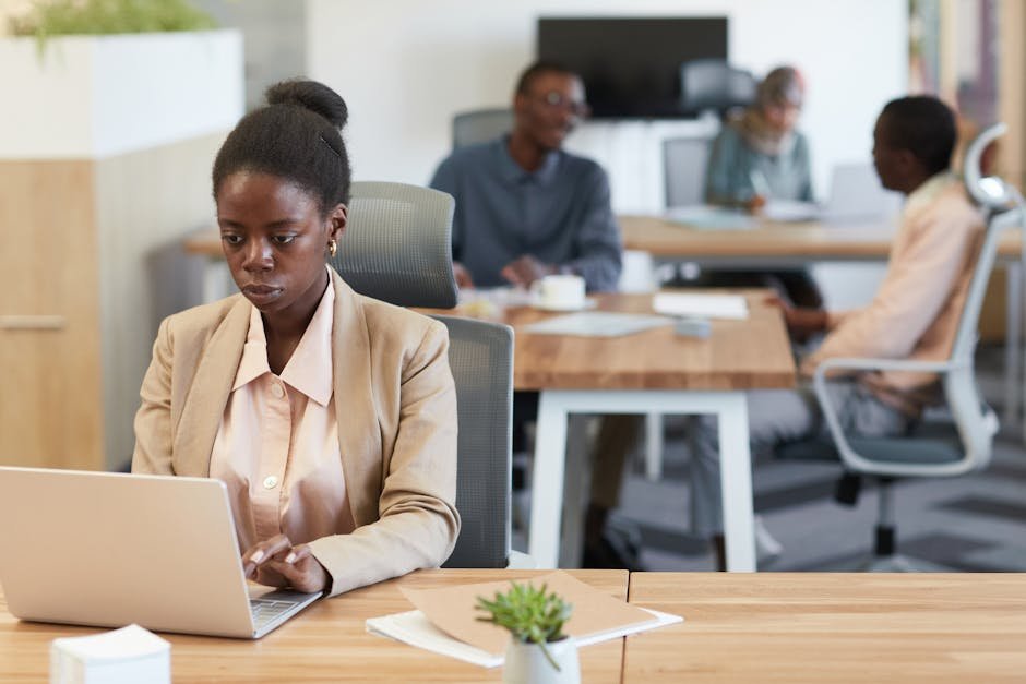 Young African woman in a blazer working intently at a laptop in a bustling office environment.