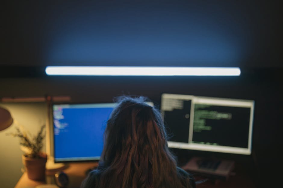 A software developer working in a dim-lit room with dual screens showing code, illuminated by a white light.