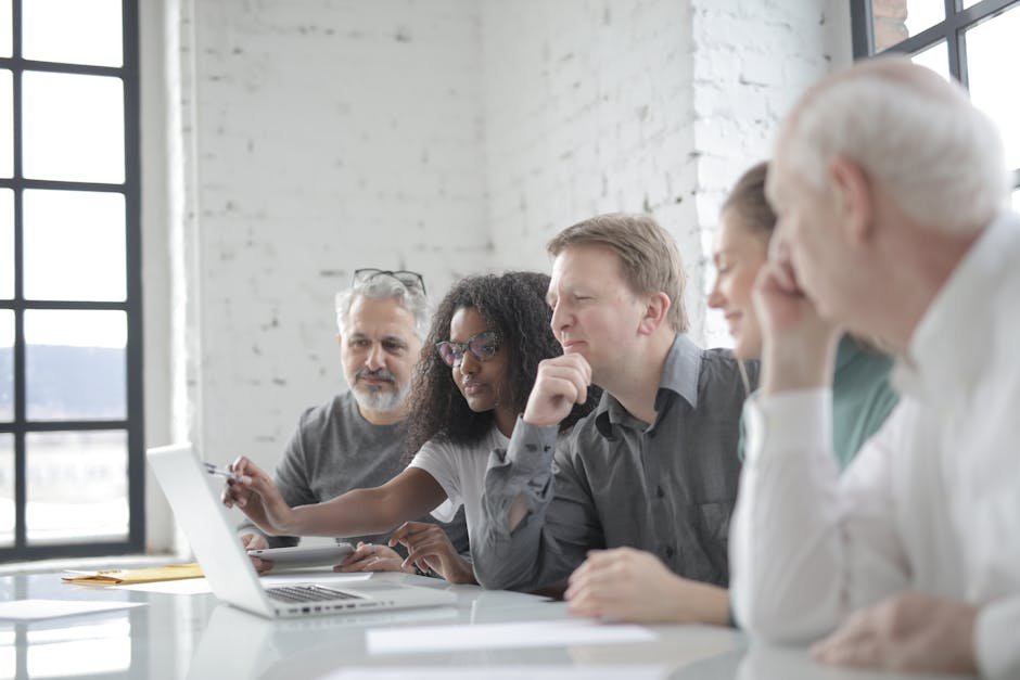 Smiling African American female worker talking about project to multiracial coworkers while pointing on netbook screen with pen and sitting at white table in light office in afternoon