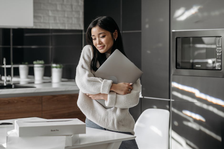 Asian woman in a cozy kitchen holding a laptop, expressing contentment.