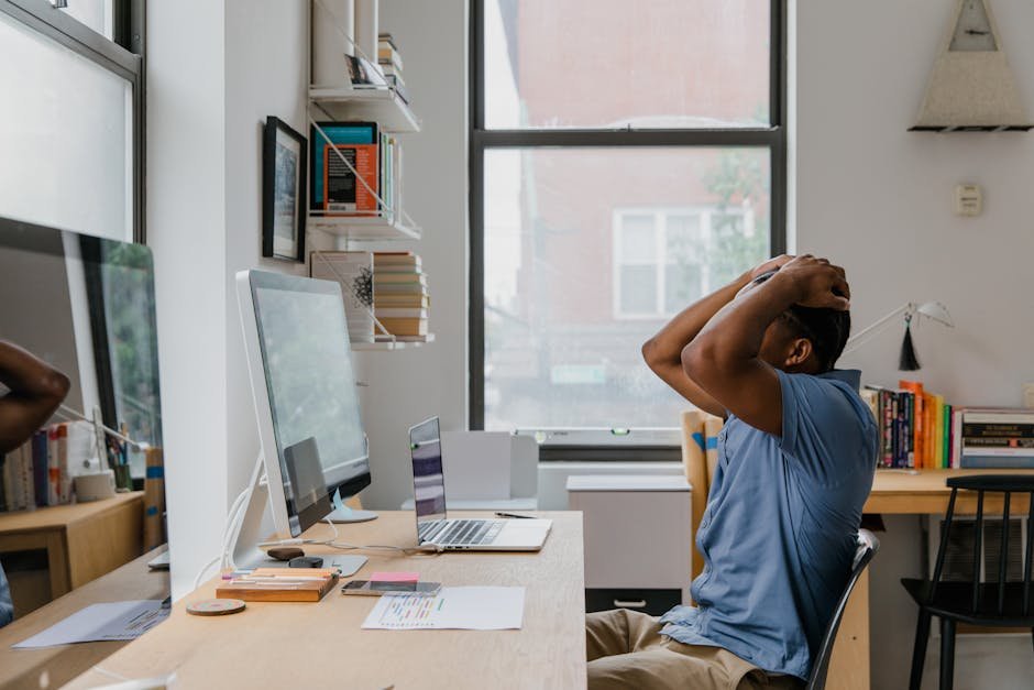 Man in casual shirt feeling stressed while working at office desk with computer and laptop.